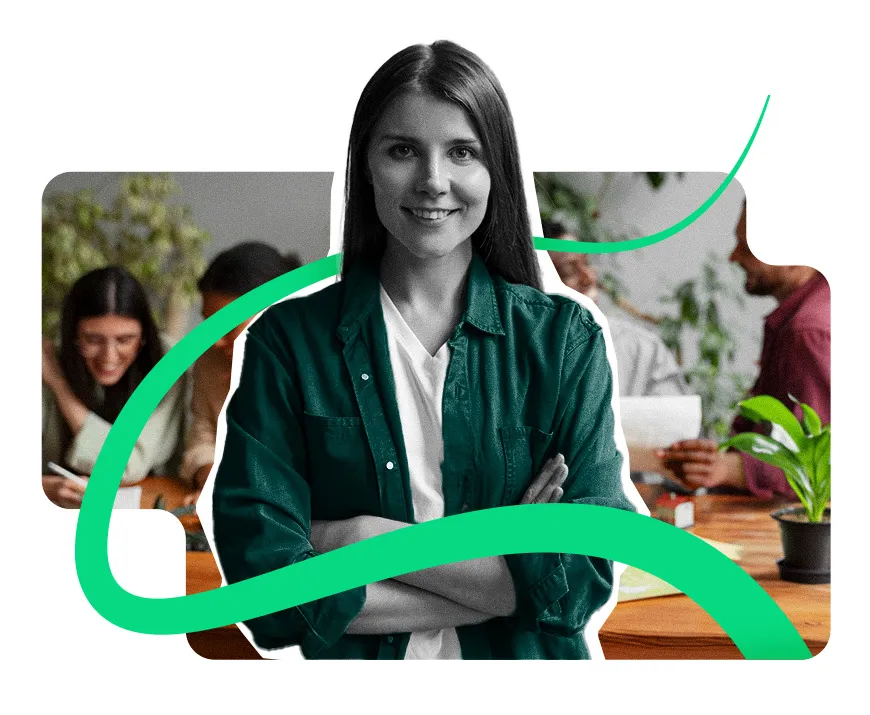 Una mujer sonriente con saco verde y los brazos cruzados, con compañeros de trabajo en una mesa detrás de ella