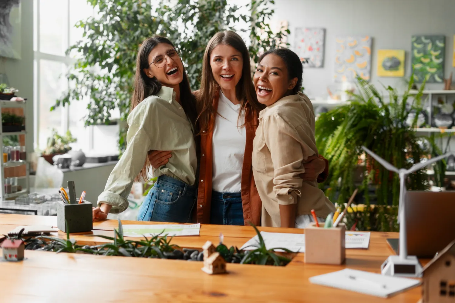 Tres mujeres abrazadas y riendo en un espacio de trabajo lleno de plantas